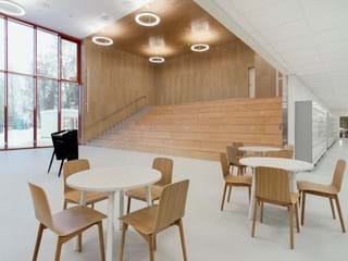 A spacious, modern interior with wooden bleacher seating against a wall, circular ceiling lights, and a mix of round tables with wooden chairs in the foreground. Large windows offer a view of a snowy scene outside.