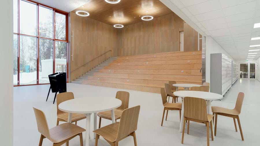 Erdvus, modernus interjeras su balintomis medinėmis sėdėjimo vietomis prie sienos, apvaliais lubų šviestuvais ir apvalių stalų ir medinių kėdžių deriniu pirmame plane. Pro didelius langus atsiveria vaizdas į sniegą lauke.