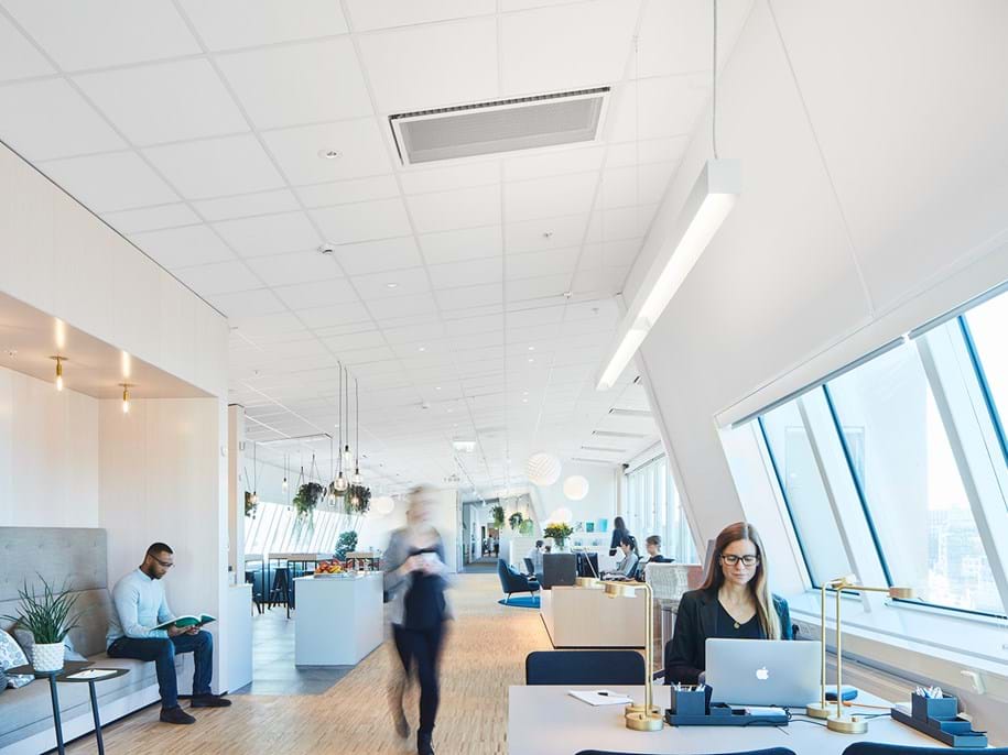 A modern, spacious office with large windows. A woman types on a laptop at a desk in the foreground. Two people sit in a lounge area, while another person walks through the space. The decor is bright and minimalist, with plants and neutral tones.