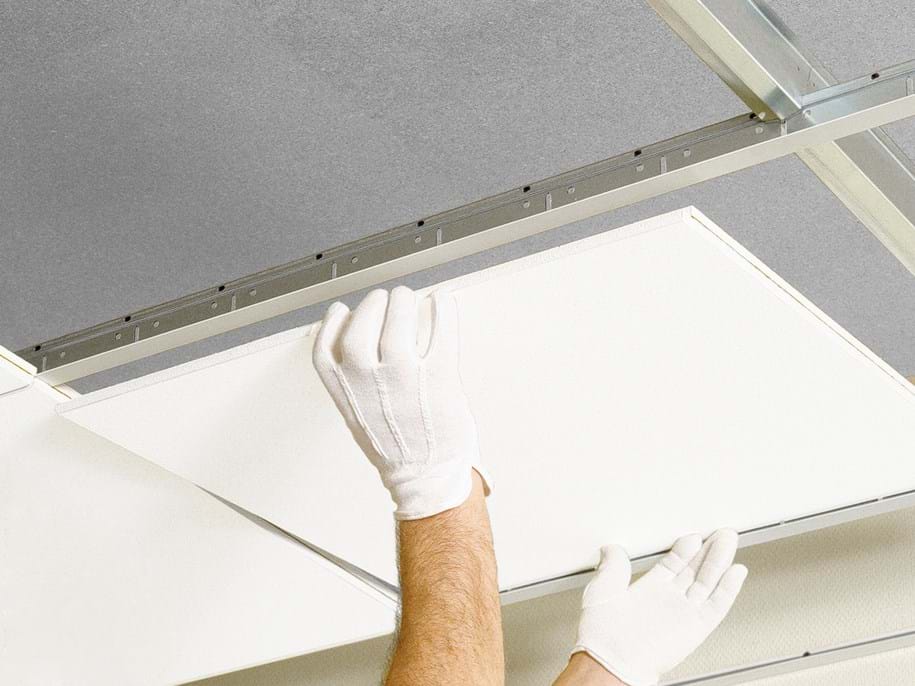 Gloved hands installing a ceiling tile into a metal grid, illustrating a suspended ceiling system. The tile is almost in place against a backdrop of other tiles and the metal framework.