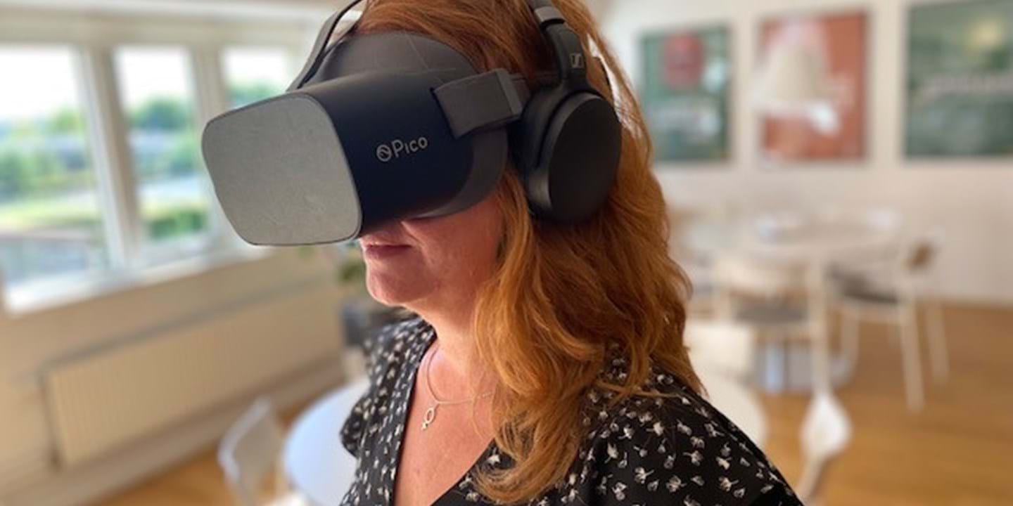 Žena s dlouhými kaštanovými vlasy má na sobě náhlavní soupravu pro virtuální realitu Pico a sluchátka. Je ve světlém, moderním pokoji s dřevěnými podlahami a bílým nábytkem. Na stěnách na pozadí se zobrazí rozmazaná umělecká díla.