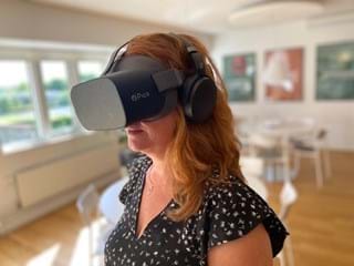 A woman with long auburn hair is wearing a Pico virtual reality headset and headphones. She is in a bright, modern room with wooden floors and white furniture. Blurred artwork is displayed on the walls in the background.