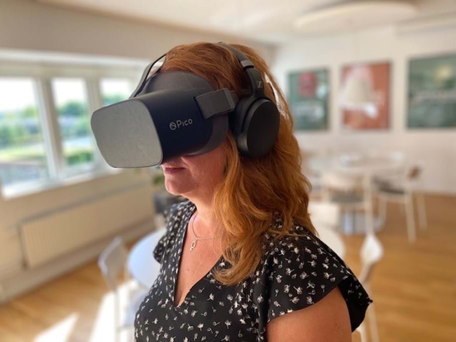 Eine Frau mit langen kastanienbraunen Haaren trägt ein Pico Virtual-Reality-Headset und Kopfhörer. Sie befindet sich in einem hellen, modernen Raum mit Holzboden und weißen Möbeln. An den Wänden im Hintergrund sind unscharfe Kunstwerke zu sehen.