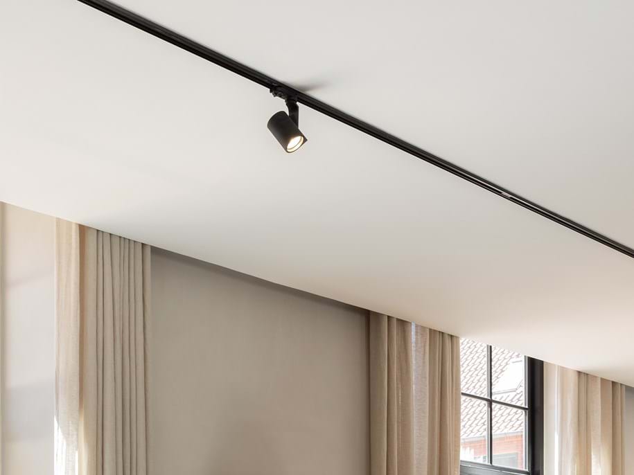Primo piano di un soffitto bianco e liscio con un binario nero per l'illuminazione. Sullo sfondo, si intravedono due finestre con tende beige.