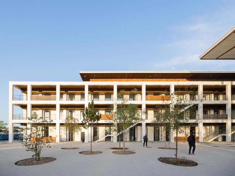 Modernes dreistöckiges Gebäude mit offenen Balkonen und einem Flachdach, das einen geräumigen Innenhof mit jungen Bäumen und ein paar Spaziergängern bietet. Der Himmel ist klar und blau und unterstreicht die moderne Architektur.