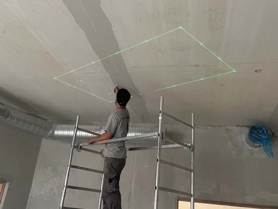 Eine Person steht auf einer Leiter und markiert mit einem grünen Laser eine rechteckige Fläche an der Decke. Die umliegenden Wände sind unbearbeitet, und in der Nähe ist ein Luftschacht zu sehen.