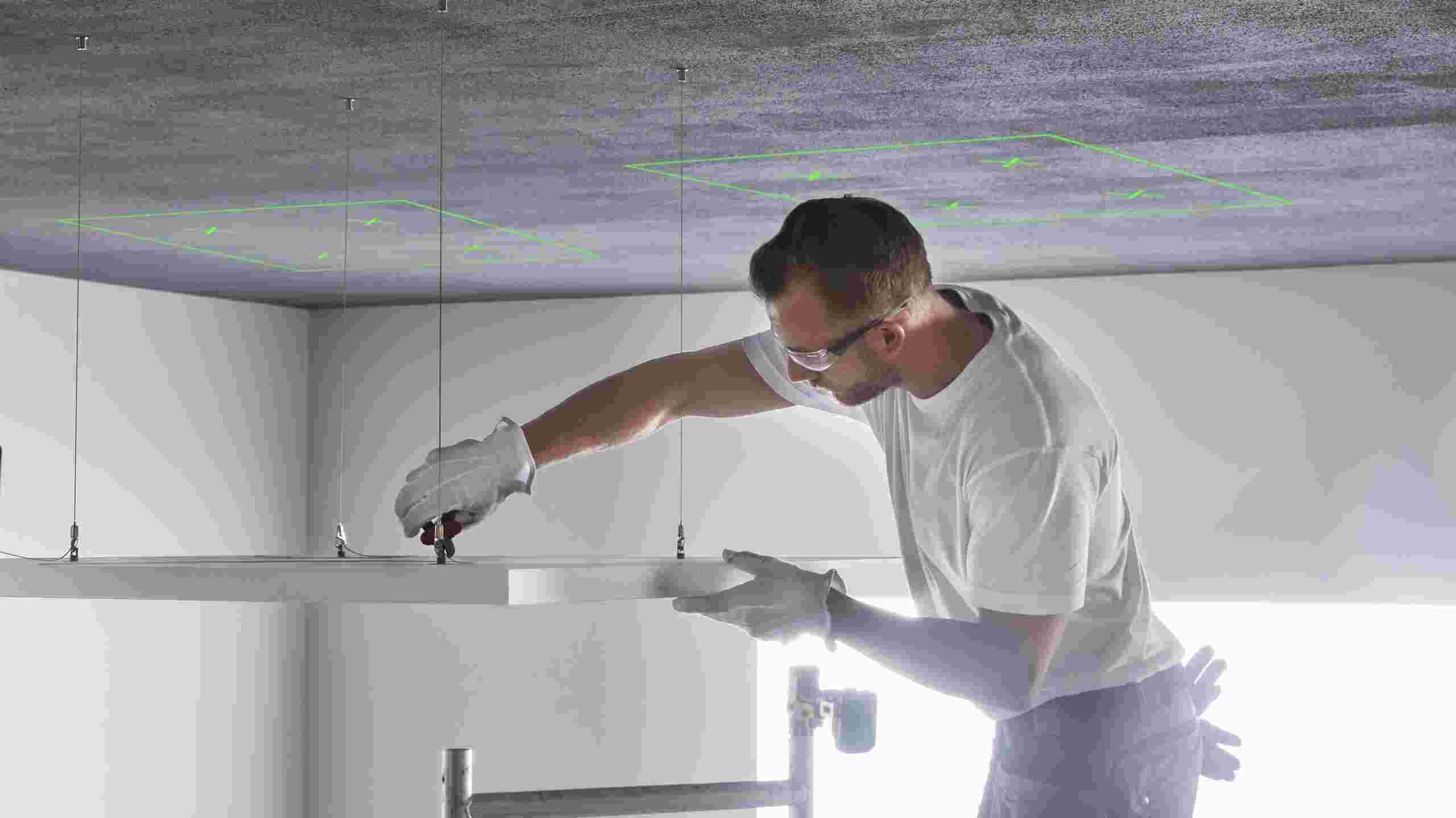 A person wearing safety glasses and gloves is installing a ceiling panel using tools. Green markings are visible on the ceiling above.