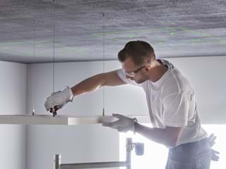 Eine Person mit Schutzbrille und Handschuhen ist dabei, eine Deckenplatte mit Werkzeugen zu montieren. An der Decke darüber sind grüne Markierungen zu sehen.