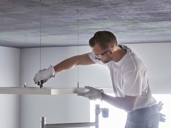 A person wearing safety glasses and gloves is installing a ceiling panel using tools. Green markings are visible on the ceiling above.