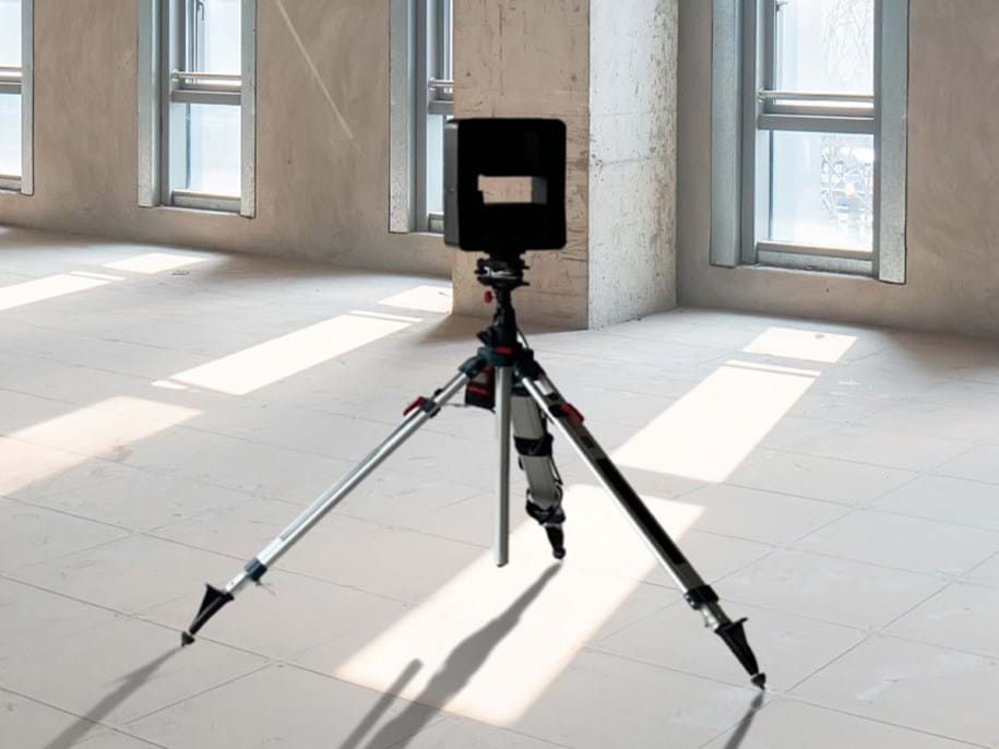 Eine Kamera auf einem Stativ steht in einem leeren Raum mit großen Fenstern. Das Sonnenlicht strömt durch die Fenster und wirft lange Schatten auf den gefliesten Boden. Der Raum wirkt industriell mit Betonwänden.