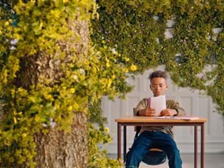 Boy reading a book at school desk in room with plants growing on the walls