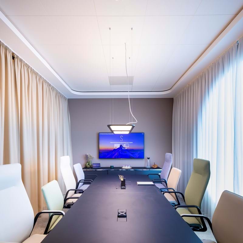 Balti, piekaramie akustiskie griesti lielā konferenču zālē ar krēsliem gaišos toņos, aizkariem logus un stikla sienām un ekrānu pie sienas