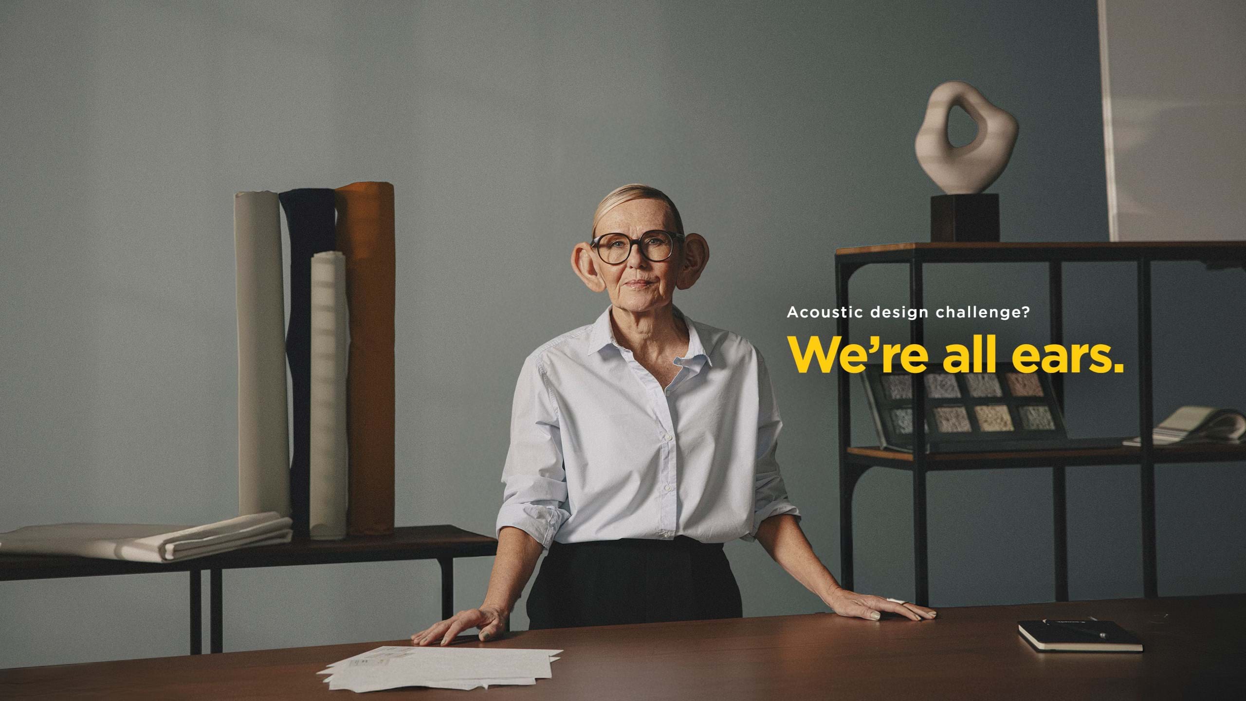 A person with oversized, cartoonish ears stands confidently at a table with acoustic design materials. The text reads, Acoustic design challenge? Were all ears. The background shows shelves with various design objects.