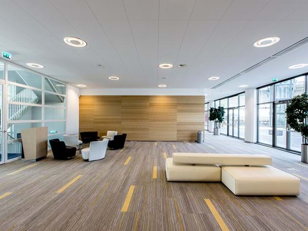 Modern office lobby with a beige sectional sofa, small table and chairs. Large windows line one wall, letting in natural light. Green potted plants are placed by the windows. The ceiling has recessed lighting, and the floor is carpeted with a striped pattern.