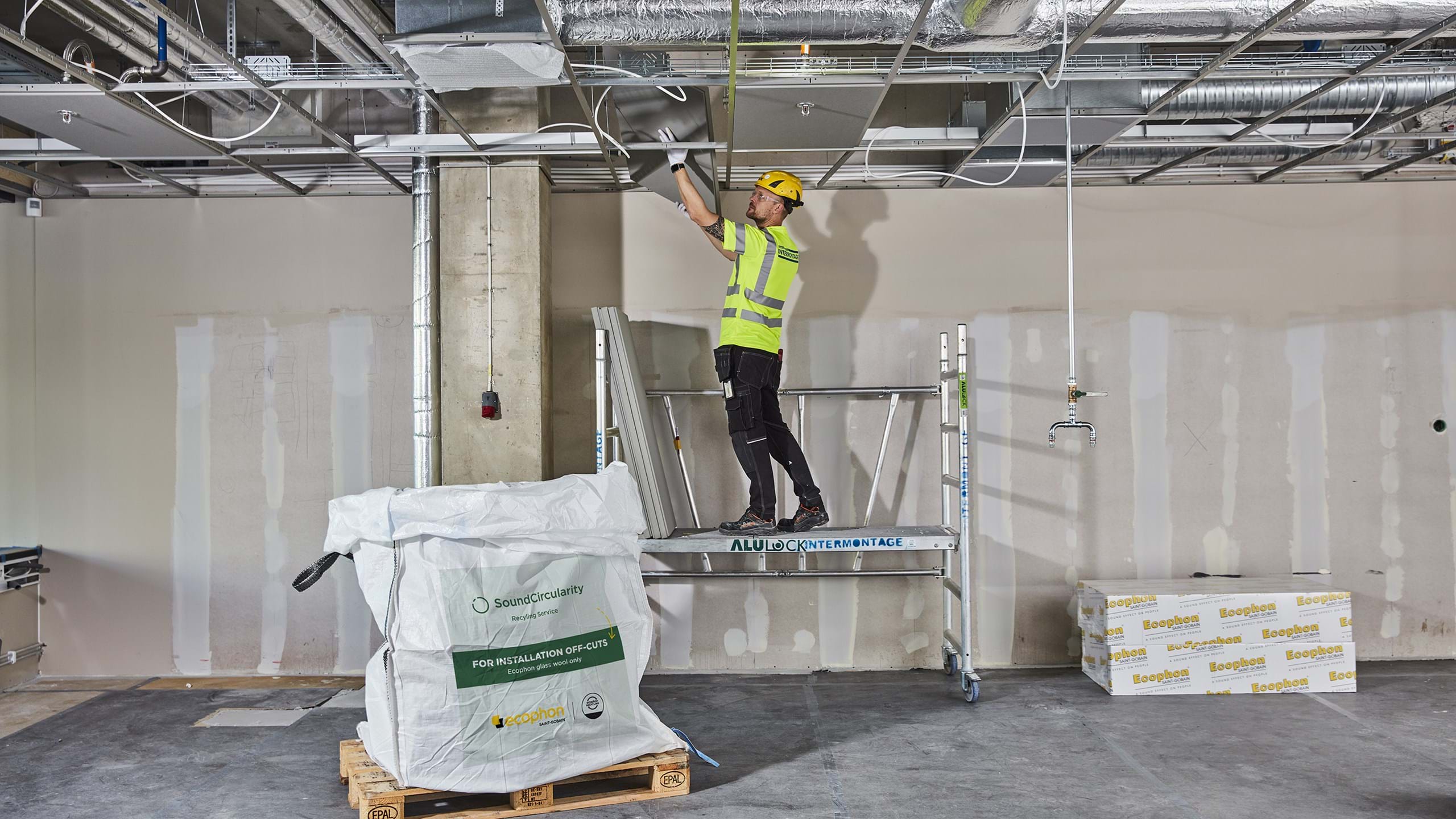 A man standing on scaffolding in an unfinished room demounting acoustic panels for recycling.