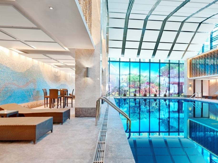 Interior of a modern building featuring an indoor swimming pool with clear blue water. The pool area has a glass wall overlooking a garden, and the ceiling is curved with structural beams. To the left, there is seating with tables and chairs on a tiled floor, adjacent to a decorative mosaic wall. The ceiling features white square and rectangular acoustic panels. The space is well-lit, creating an inviting and luxurious atmosphere.
