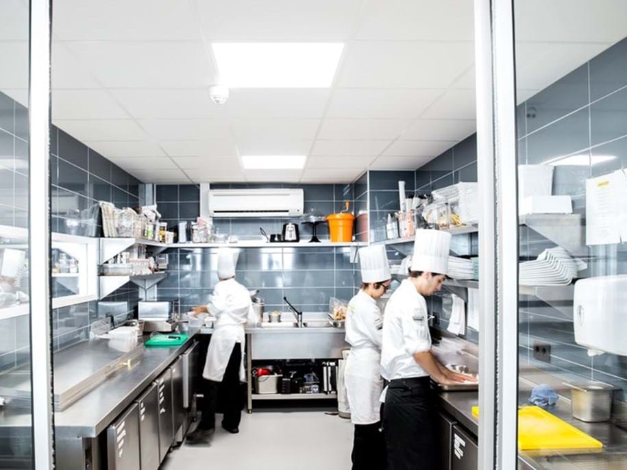 A professional kitchen with stainless steel appliances and surfaces. Two chefs in white uniforms and chef hats are working in the kitchen. One chef is focused on preparing food at a stainless steel counter, while the other is cooking at a stove. The kitchen is clean and well-organised, with various pots, pans, and kitchen utensils visible on shelves and counters. A white acoustic ceiling including lighting is featured. The image conveys a busy yet orderly culinary environment.