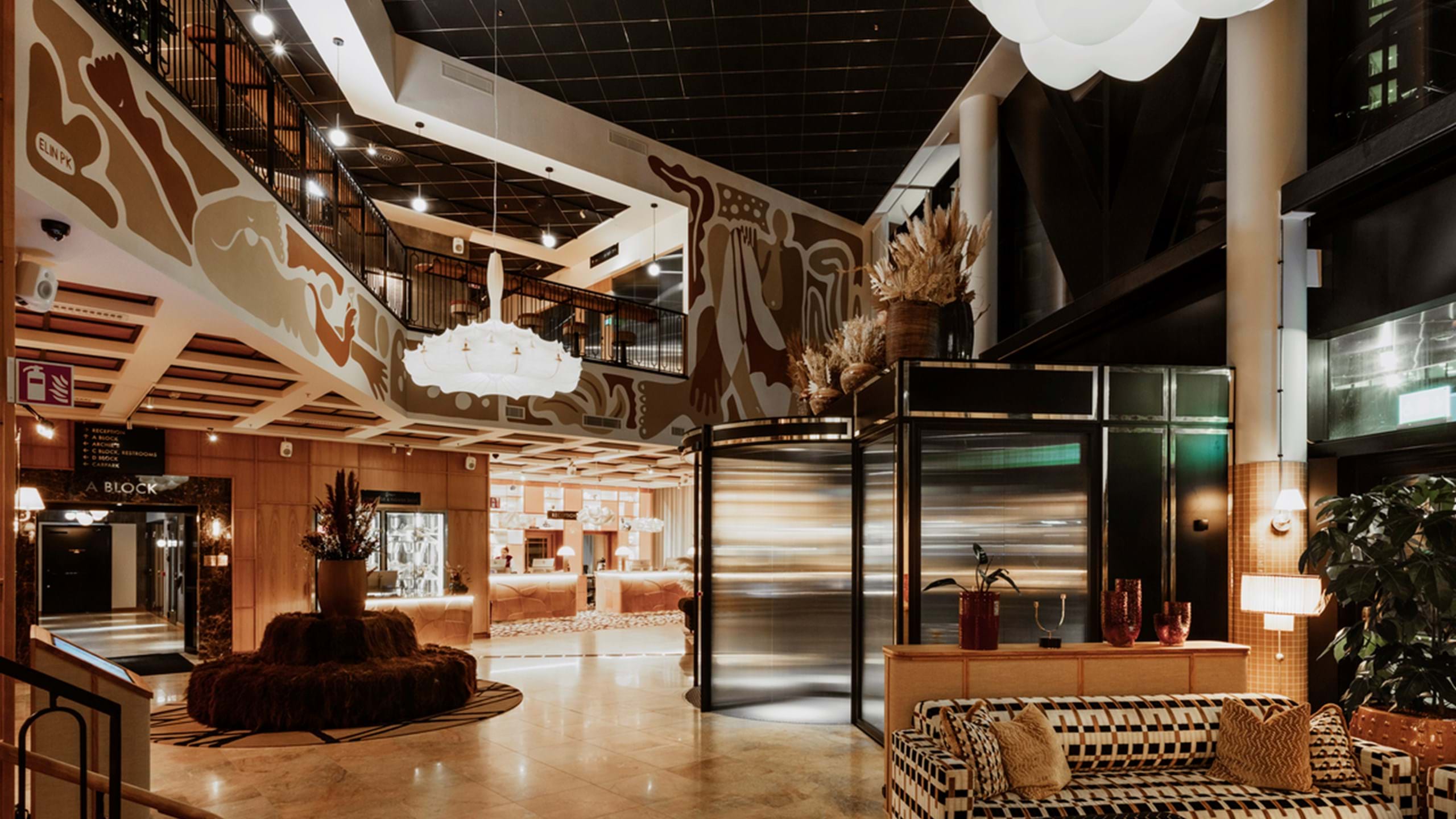 Modern hotel lobby with warm ambient lighting. The space features a mix of contemporary and art deco design elements, including a patterned acoustic ceiling, decorative wall panels with abstract designs, and stylish furniture. A central seating area with plush sofas and chairs is arranged around a circular wooden table adorned with large feathered plants. To the right, there is an elevator with reflective doors next to potted plants. The lobby extends into the background where more seating areas and hotel amenities are visible under soft, glowing light fixtures that resemble clouds.