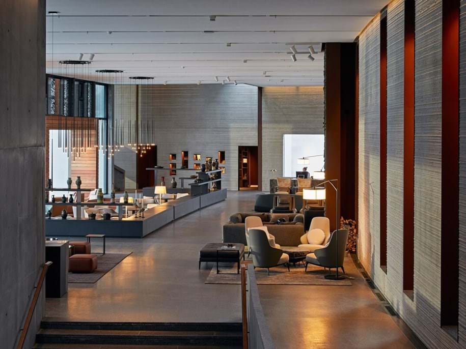 A modern hotel lobby with minimalist design, featuring sleek furniture, warm lighting, and polished concrete floors. The space includes seating areas with cushioned chairs, a long reception desk, and elegant pendant lights, all in a warm, neutral color palette. The ceiling is covered with white acoustic fabric.