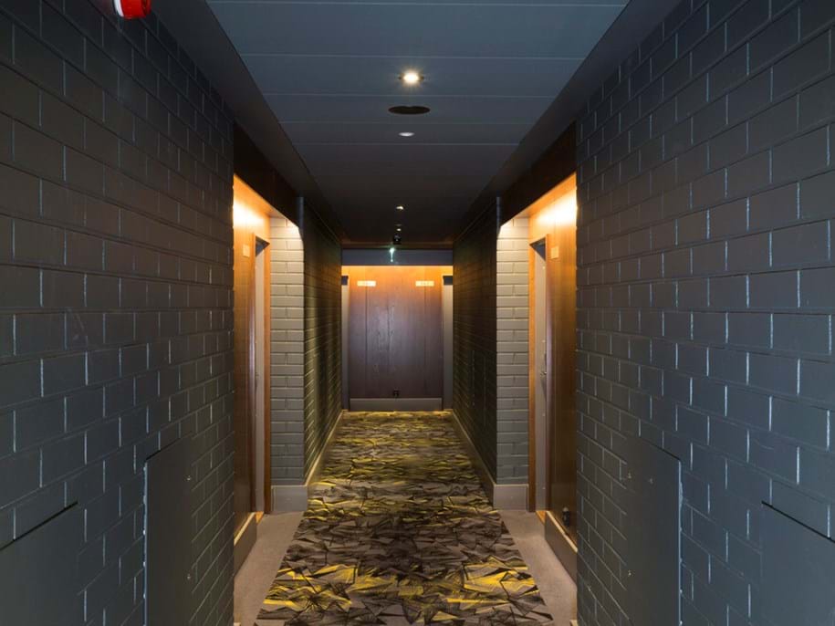 A narrow corridor with dark grey brick walls and a patterned carpet with leaf designs. The corridor is lit by recessed ceiling lights and wall-mounted fixtures at each door, creating a warm ambiance. An acoustic ceiling is featured throughout the corridor. At the end of the hallway, there appears to be an elevator with its doors closed.