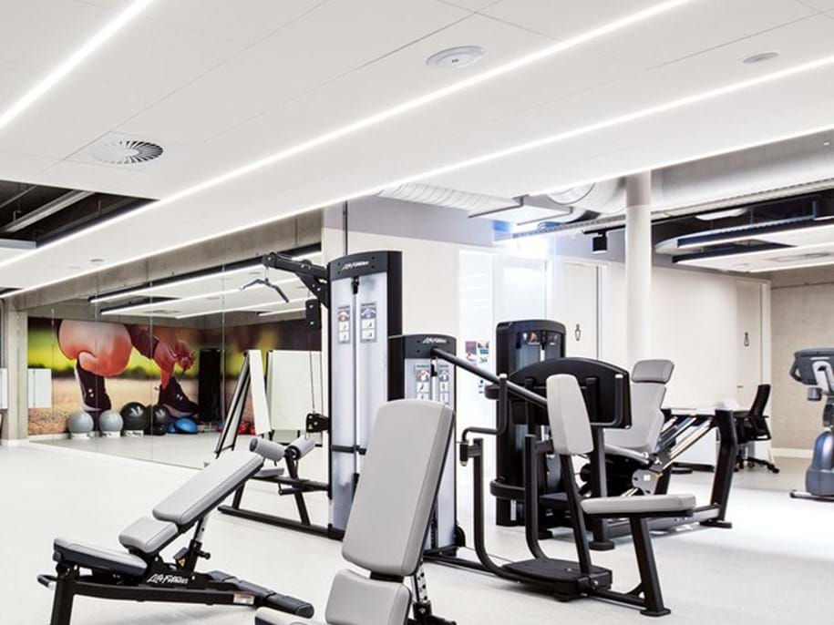 A modern gym interior with various exercise equipment. The space is well-lit with strip lighting on the acoustic ceiling, creating a bright and clean atmosphere. In the foreground, there are weight training machines and benches, while in the background, cardio machines like treadmills are visible. The walls are adorned with large images of fruits, suggesting a focus on health and nutrition.