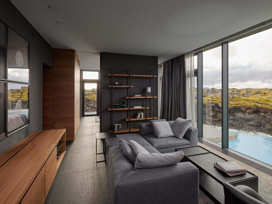 A modern living room with large floor-to-ceiling windows offering a view of a natural rocky landscape and a pool. The room features gray sofas, a wooden cabinet, and a wall-mounted bookshelf. The interior has a minimalist design with dark walls, simple furniture, and natural light streaming in through the windows. The ceiling is covered with grey acoustic fabric.