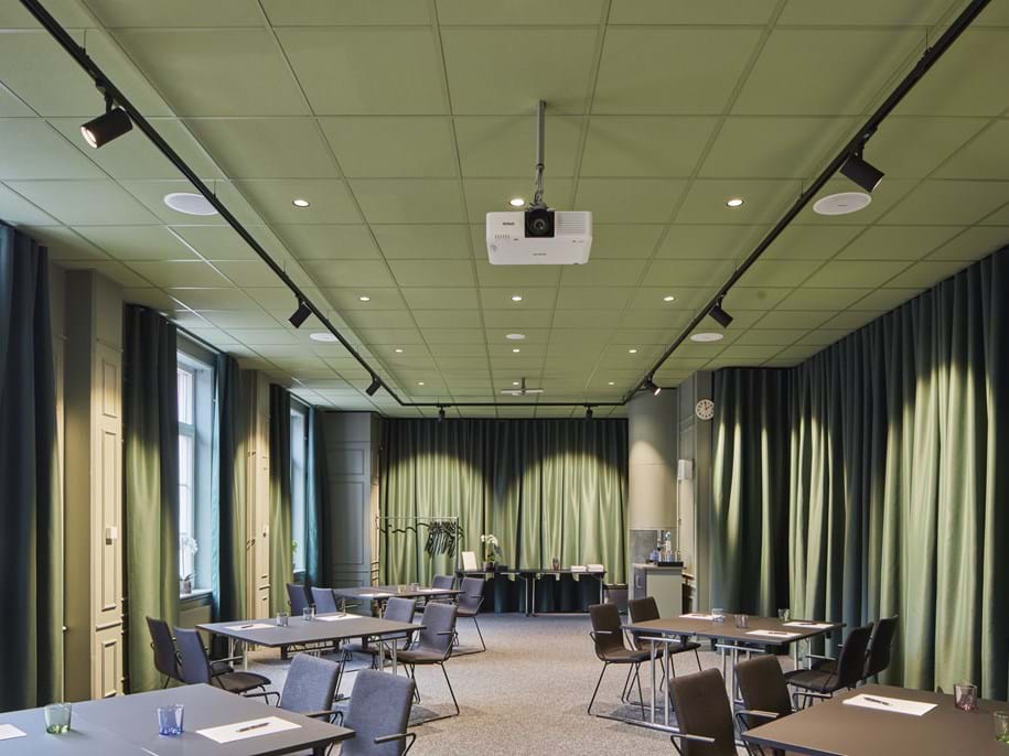 A modern conference room with green walls and acoustic ceiling, featuring several tables and chairs arranged for a meeting. A projector is mounted on the ceiling, and large windows with green curtains line one side of the room. The space is well-lit with a combination of recessed and track lighting.
